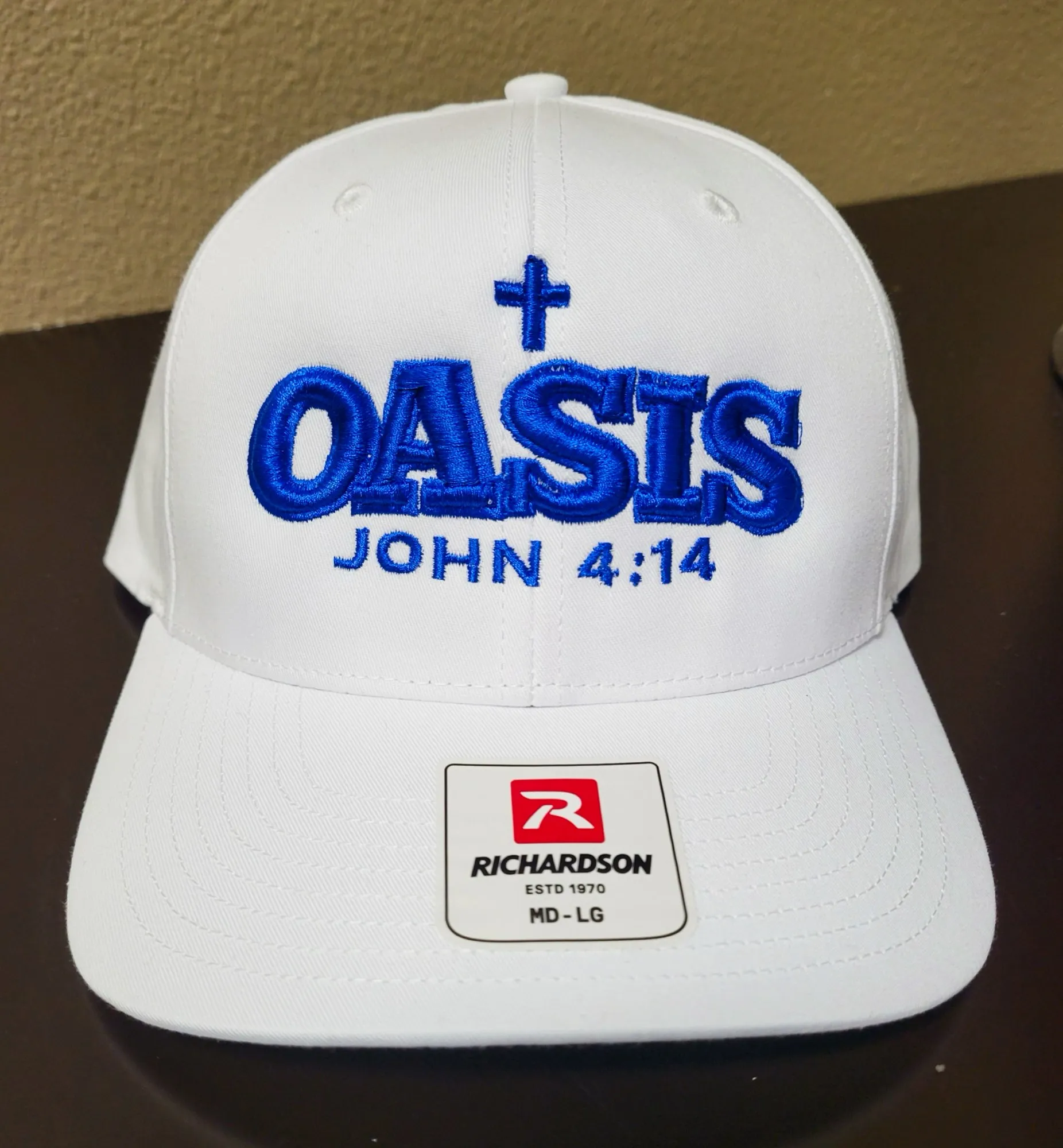 White cap with raised royal blue embroidery reading OASIS, John 4:14, with a cross above