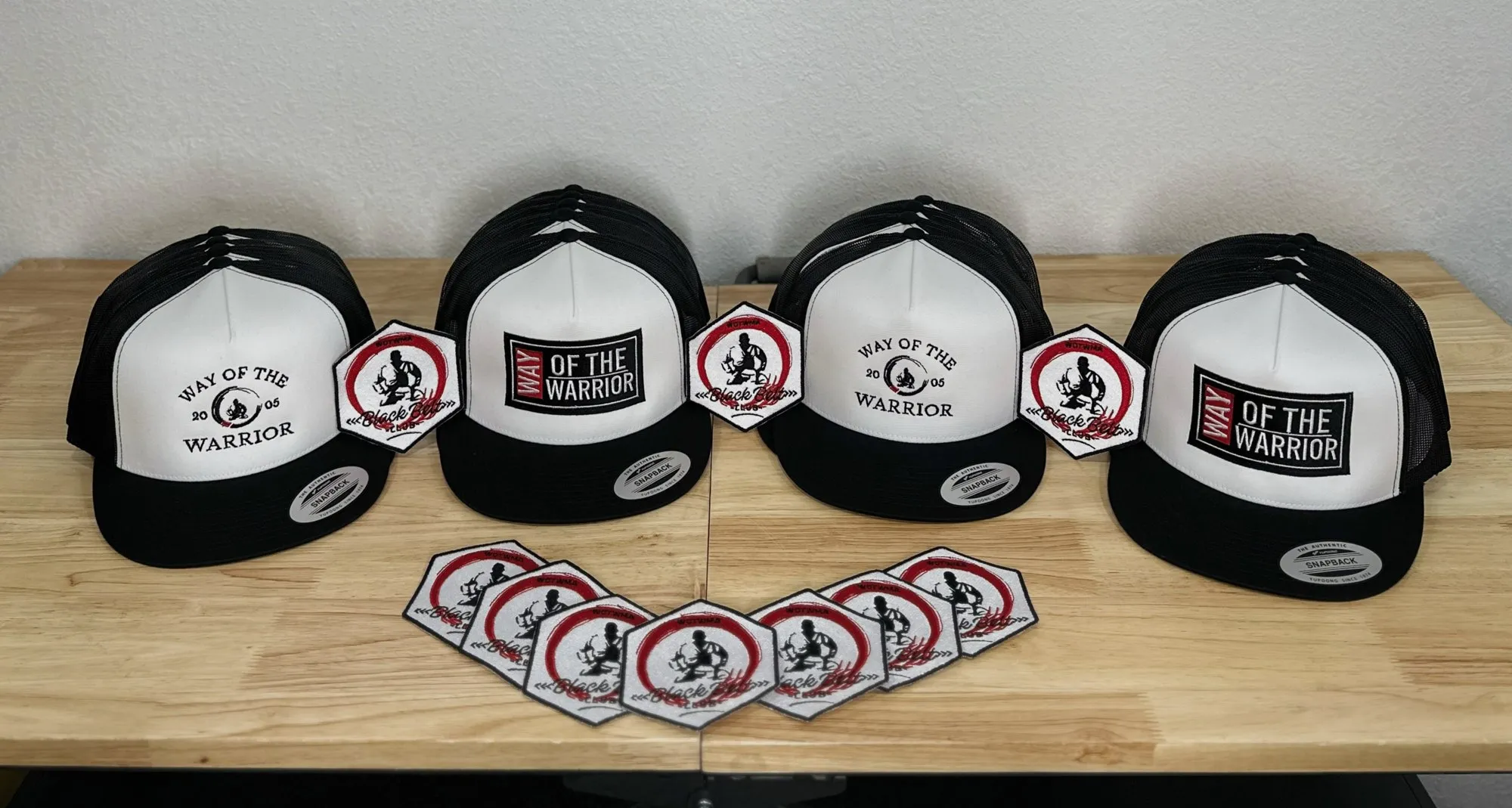 Four trucker and snapback hats with Way of the Warrior logos, set above a row of embroidered hexagon patches