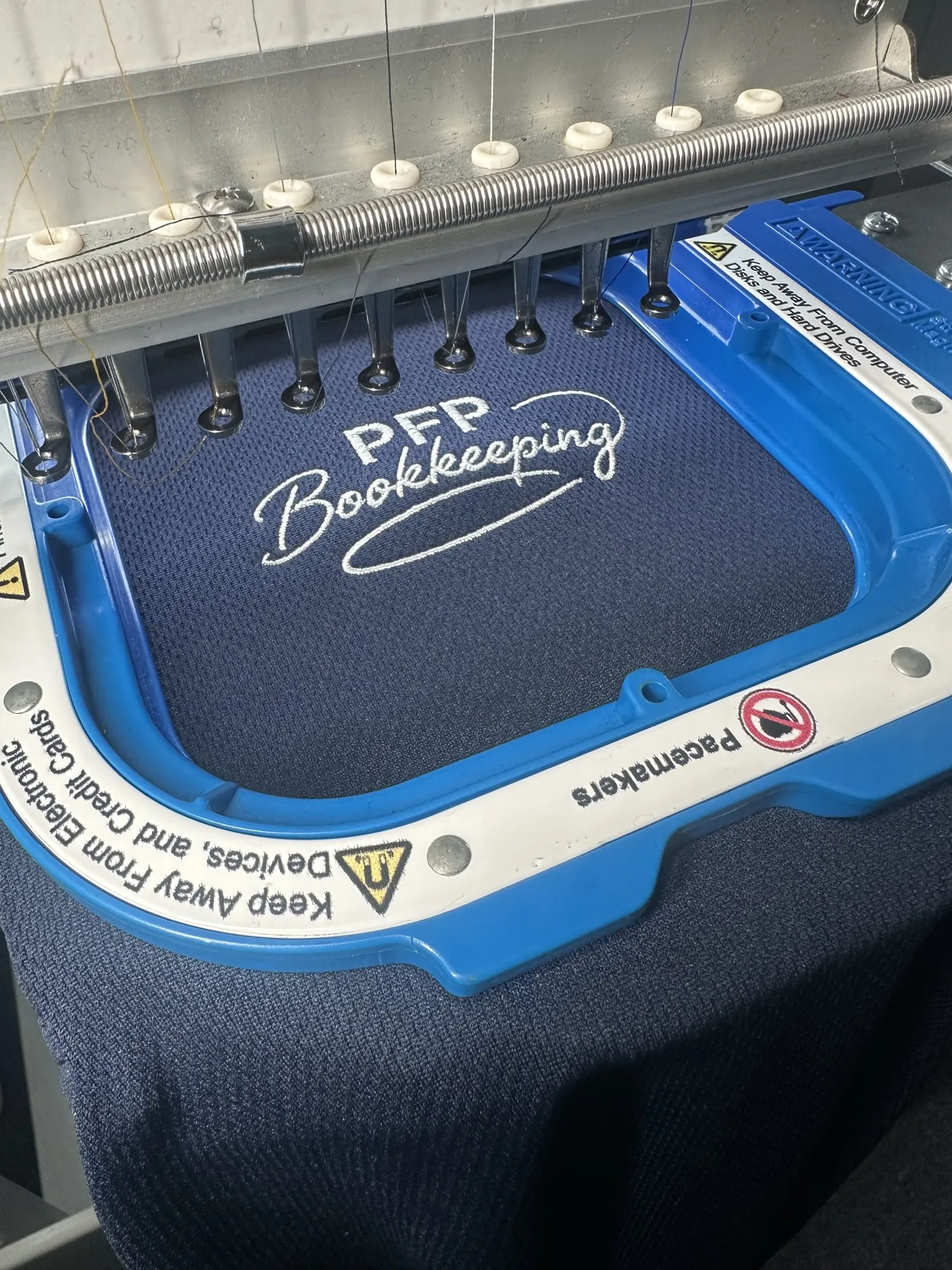 Close-up of an embroidery hoop mid-stitch, white thread spelling PFP Bookkeeping on navy fabric