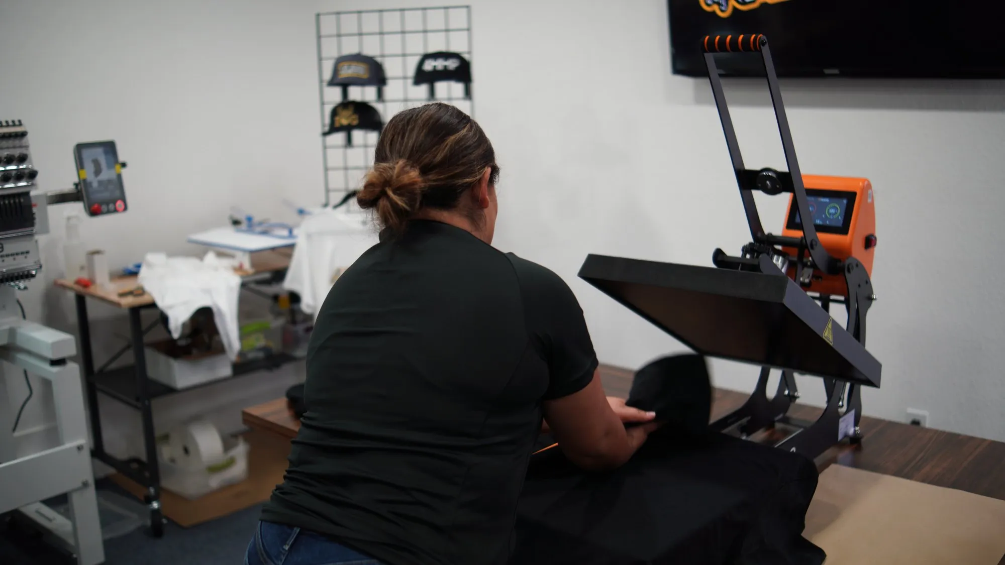 A 4 His Highest Promotions team member loading a black shirt into a heat press in the shop, with a hat wall and embroidery machine behind her